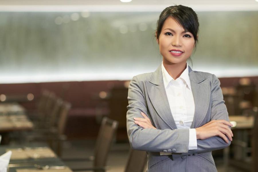 A professional portrait of a woman in a grey blazer standing with arms crossed in a formal restaurant or banquet hall.