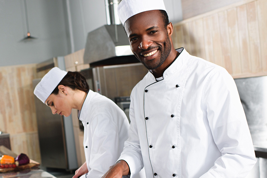 A smiling chef in a white coat and hat stands in a professional kitchen while a colleague works at a prep station in the background.