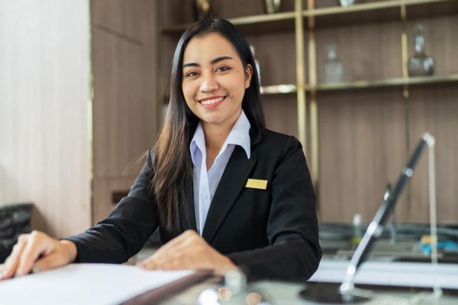 A smiling woman in a black professional blazer and name tag sitting at a front desk in a luxury hospitality or hotel setting.