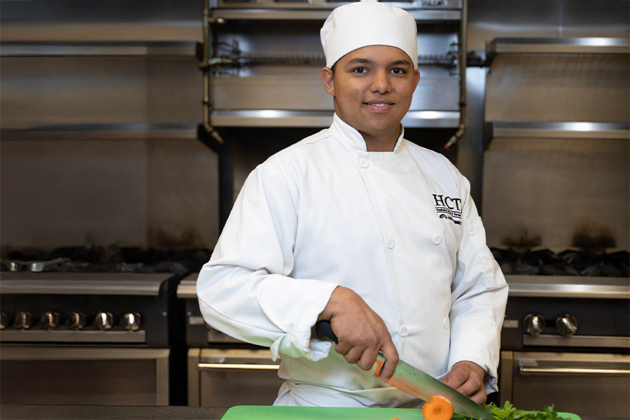 A culinary student in a white chef's coat and hat smiles while chopping vegetables on a green cutting board in a professional kitchen with commercial ranges in the background.