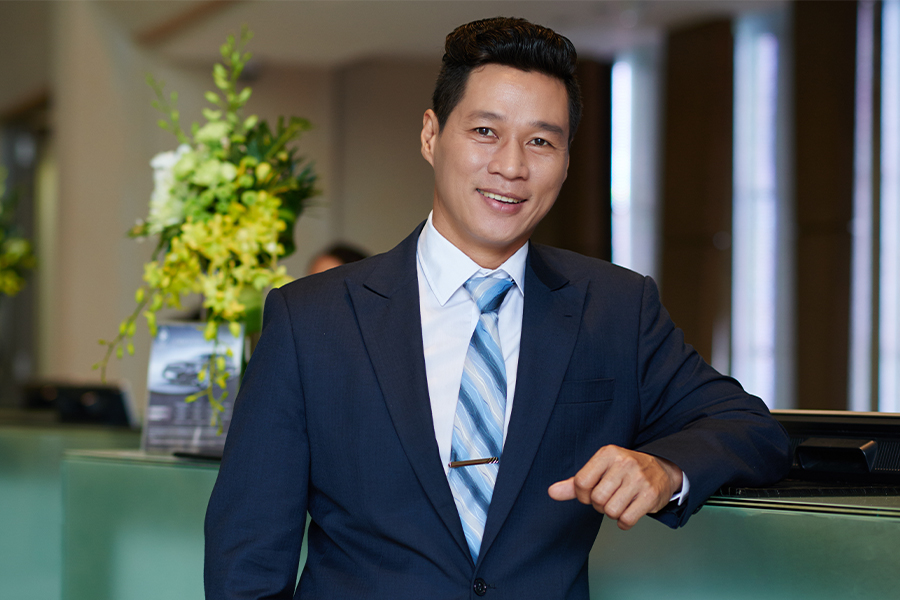 A smiling hospitality manager in a navy suit and blue striped tie leaning confidently against a front desk counter in a modern hotel lobby.
