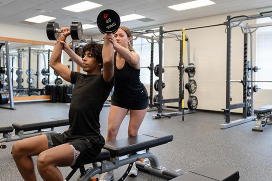 A female personal trainer assists a male athlete with a seated dumbbell shoulder press in a bright, modern gym. She provides a steady spot as he lifts weights while seated on a workout bench.