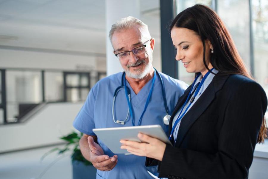 A healthcare professional in blue scrubs and a professional administrator reviewing information on a tablet.