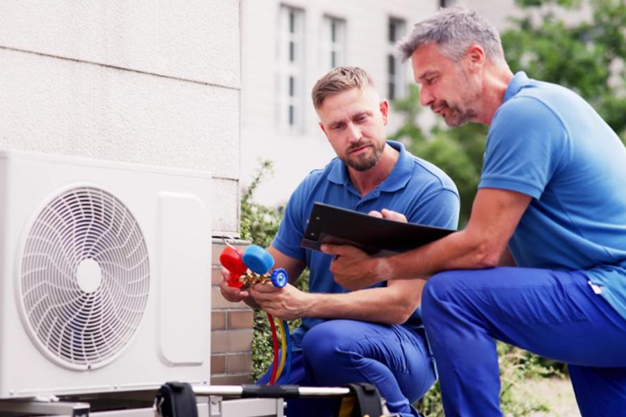 Two HVAC technicians in blue uniforms perform maintenance on an outdoor white air conditioning condenser unit. One technician holds a manifold gauge set with red and blue dials, while the other reviews a clipboard, both crouched beside the unit against a brick and light-colored wall.