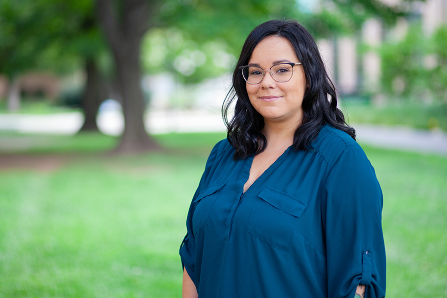 A woman with dark, shoulder-length wavy hair and glasses smiles warmly while wearing a teal blouse, standing in front of a soft-focus background of green trees and a campus lawn.
