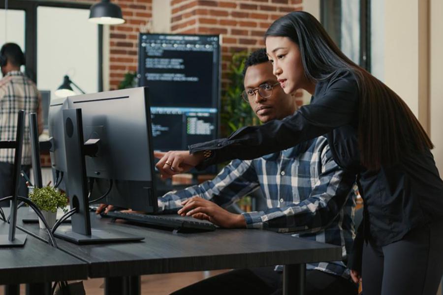 Two young IT professionals, a man and a woman, collaborate at a dual-monitor workstation in a modern office, reviewing lines of code on the screen.