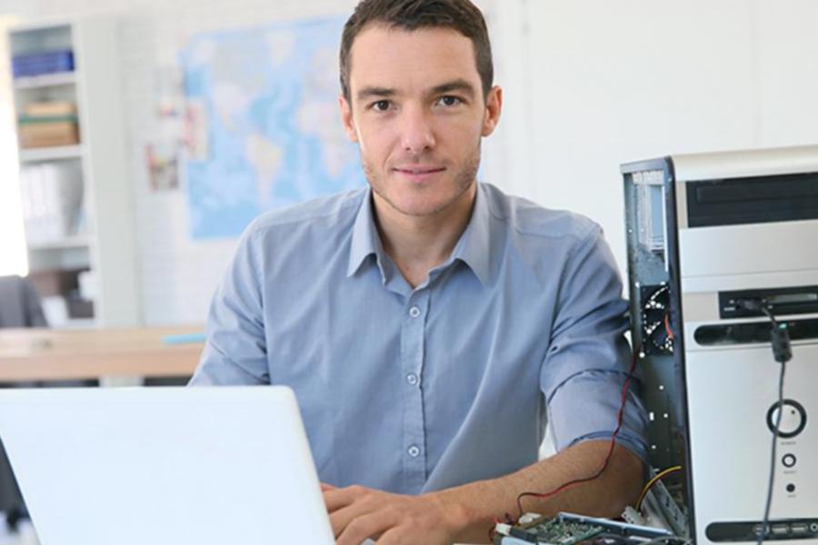 A male IT professional or student working on a laptop at a desk with a partially disassembled computer tower next to him.
