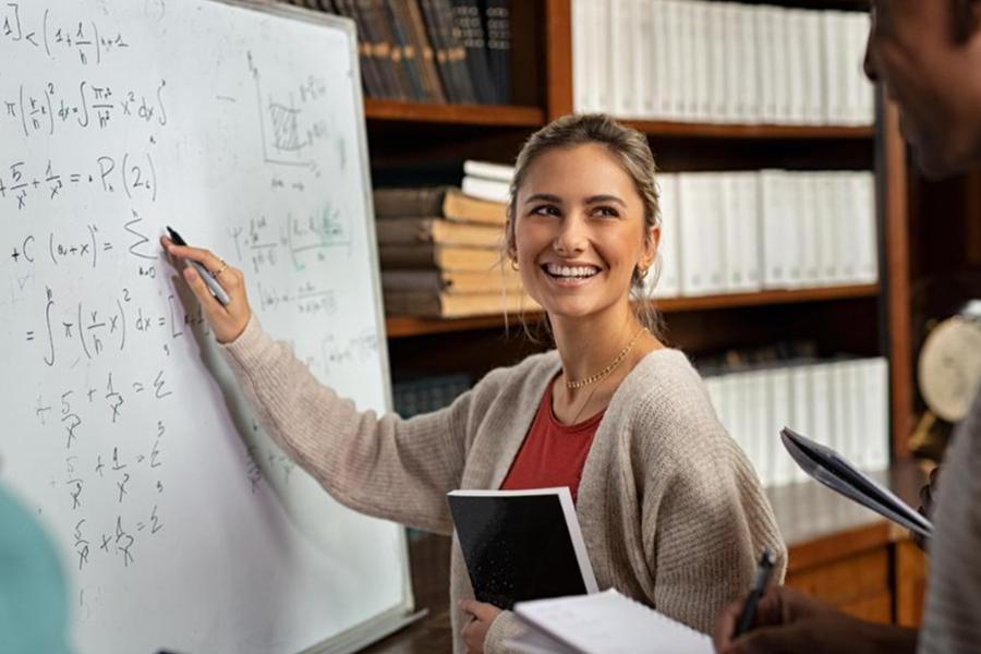 A smiling student in a tan cardigan writes complex calculus equations on a whiteboard during a group study session in a library.