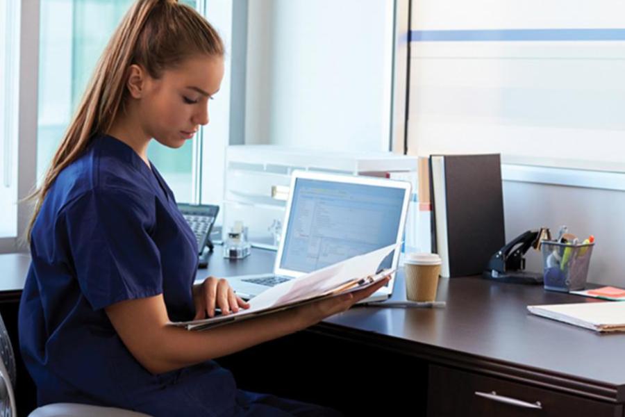 Healthcare professional in navy scrubs reviewing patient records at a desk with a laptop in a medical office setting