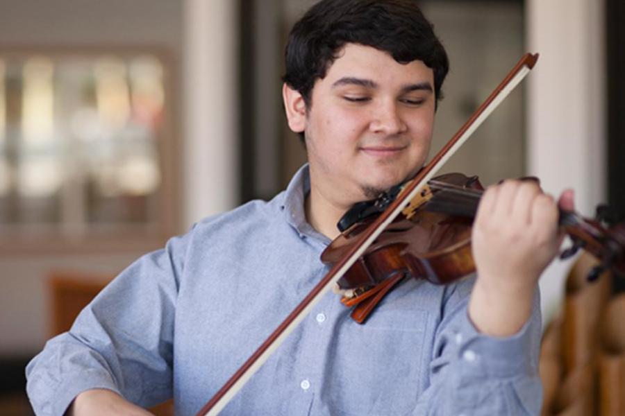 A young man in a light blue button-down shirt smiles as he plays the violin indoors.