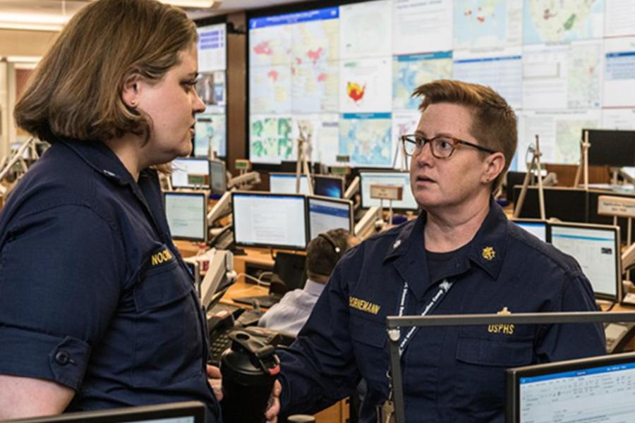Two women in navy USPHS uniforms discussing work in a high-tech operations center.