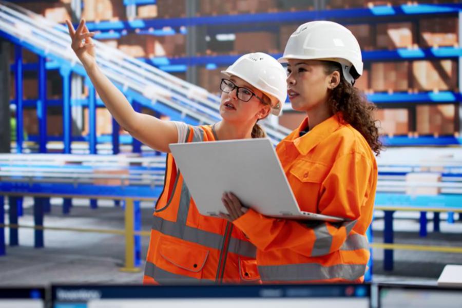 Two female engineering or logistics students in orange safety gear and white hard hats use a laptop to analyze operations in a high-tech automated warehouse.