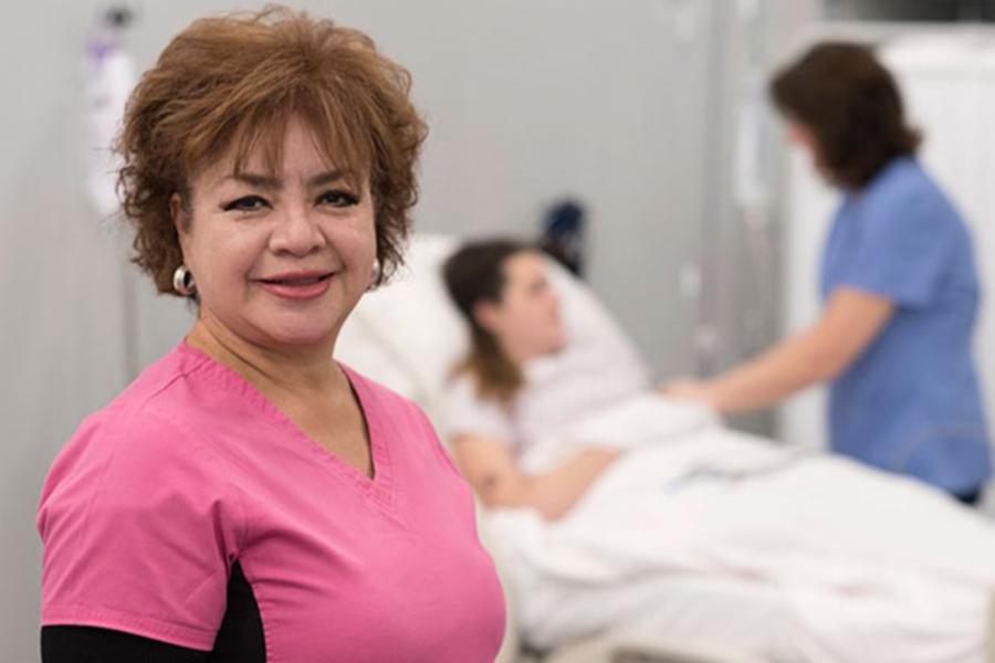A nursing assistant in pink scrubs smiles in a clinical care setting.