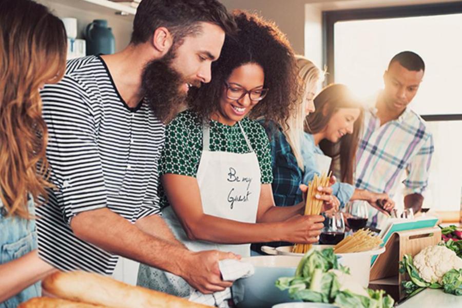 A diverse group of students smiling and preparing a pasta meal together in a bright kitchen.