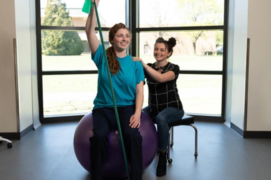 A physical therapy student assists a patient seated on an exercise ball with resistance band stretches in a bright clinical studio.