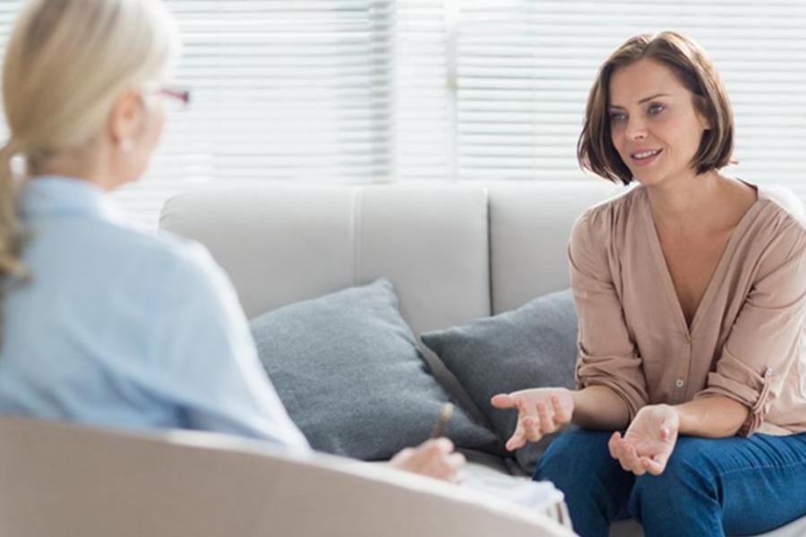 A mental health counselor or social worker in a light blue shirt listens attentively while taking notes as a female client speaks during a therapy session on a comfortable grey sofa.