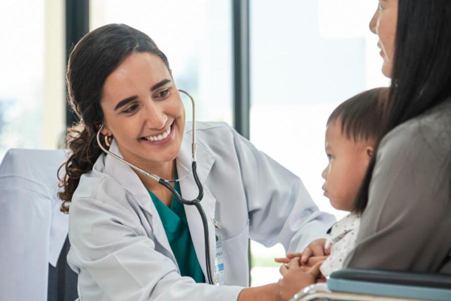 A smiling female doctor in a white lab coat uses a stethoscope to examine a young child sitting on a parent's lap.