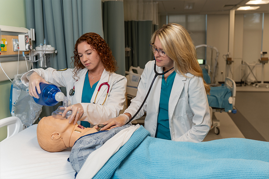 Two medical students in white lab coats and teal scrubs perform emergency resuscitation and cardiac monitoring on a patient mannequin in a simulated hospital ward.