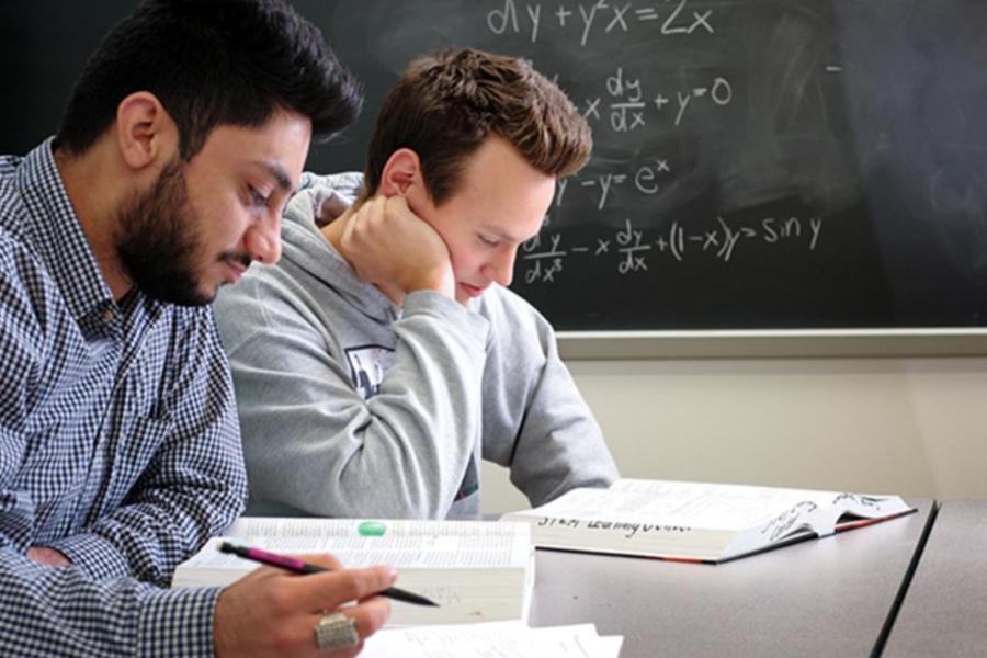 Two college students studying advanced mathematics from textbooks in a classroom with a chalkboard full of calculus equations.