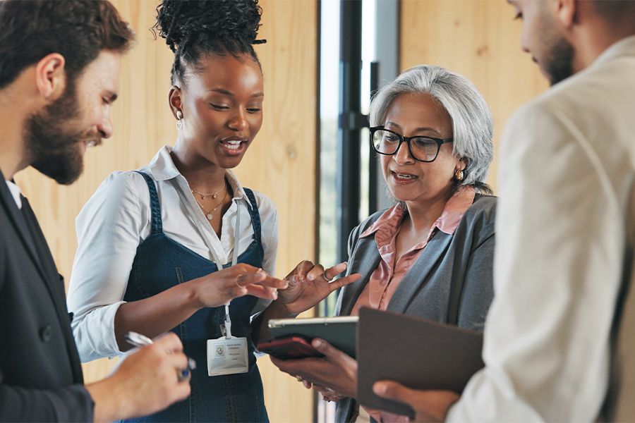Four professionals gather in discussion, with a young woman holding a tablet gesturing as she speaks to an older woman with glasses. Two colleagues look on and take notes.