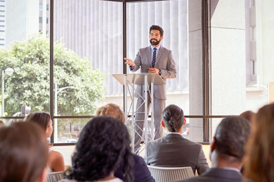 A male professional in a grey suit gives a presentation from a podium to an audience in a bright, modern conference room with floor-to-ceiling windows.