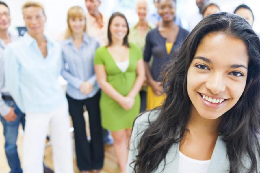 A close-up portrait of a smiling young woman with long dark hair, with a diverse group of students or colleagues blurred in the background.