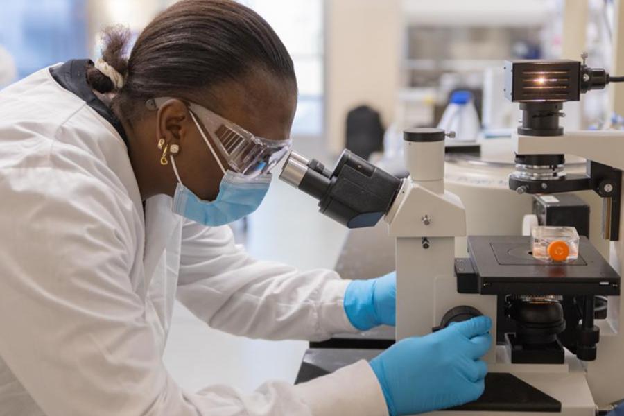 A scientist in a lab coat, mask, and safety goggles examines a sample through a microscope.