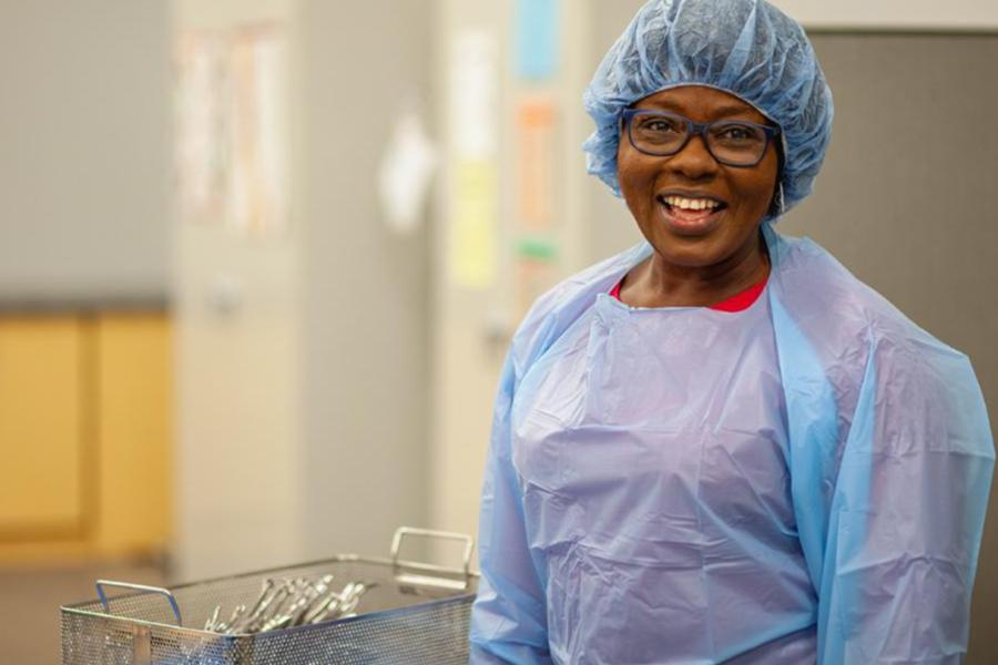 A sterile processing technician in a blue gown and hair cover smiles while holding a metal instrument tray in a healthcare facility.