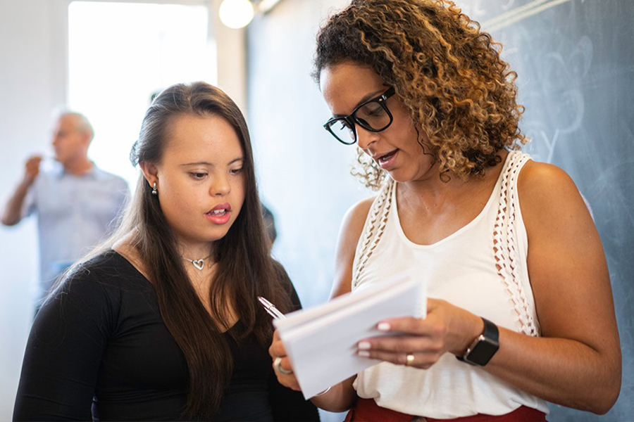 A student with Down syndrome and a woman wearing glasses review papers together in a classroom setting, with other people visible in the background near a chalkboard