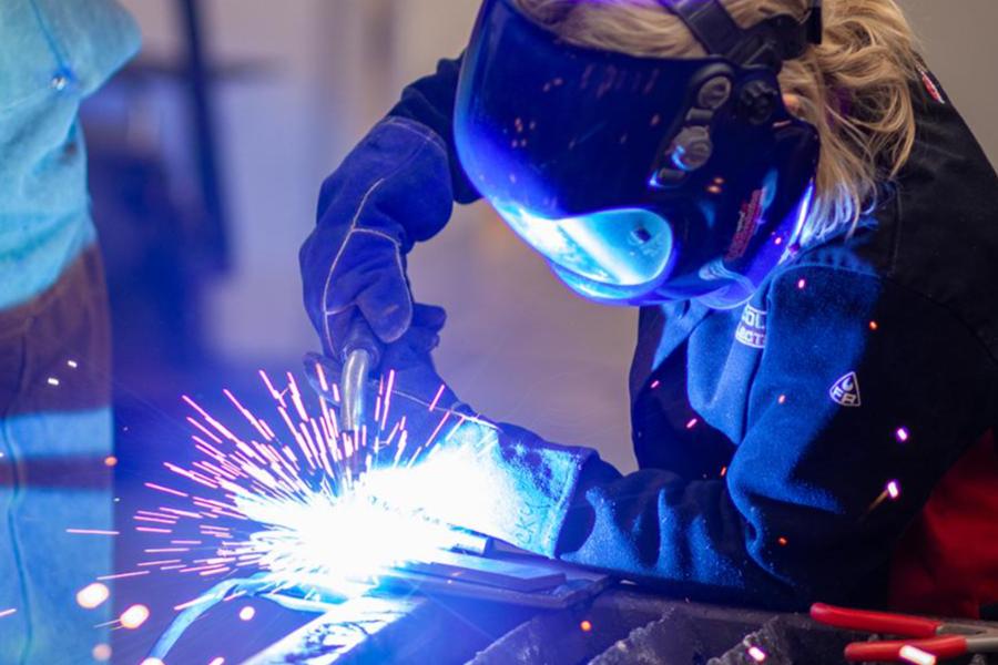 A welder wearing a protective helmet and gloves works on a metal piece, with bright sparks flying from the weld point.