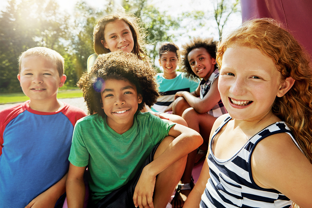 A group of six children smile together outdoors on a sunny day, with trees and bright sunlight visible in the background.