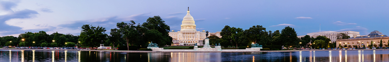 A panoramic twilight view of the U.S. Capitol building reflected in the Capitol Reflecting Pool, surrounded by illuminated trees and monuments under a blue evening sky.