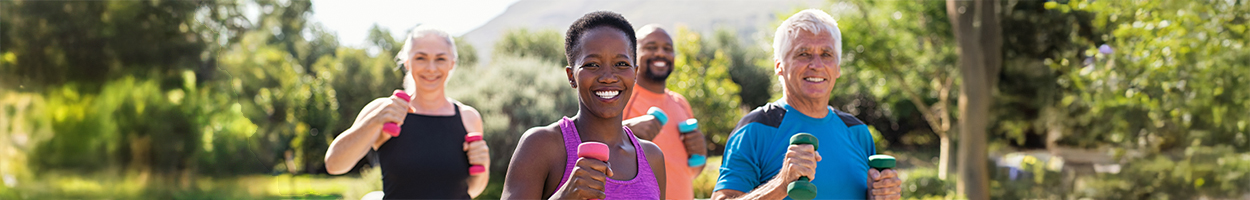 A diverse group of four smiling adults exercise outdoors with hand weights in a sunny, tree-lined setting with mountains visible in the background.