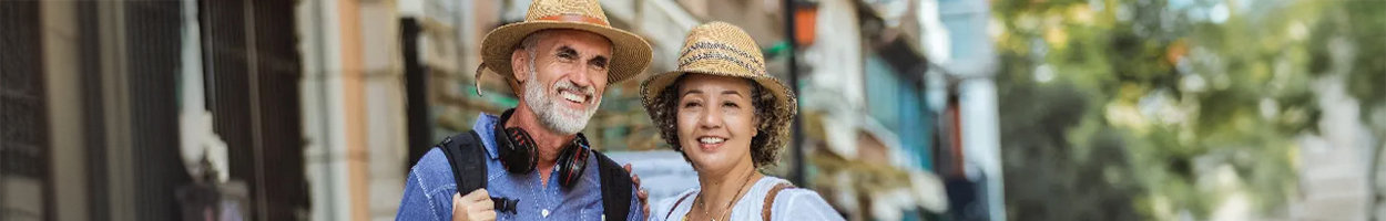 Two smiling middle-aged adults wearing straw hats pose together on a sunny street, with the man carrying a backpack and headphones around his neck.