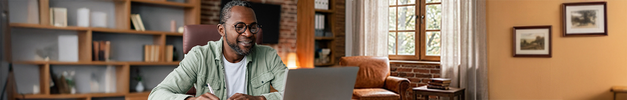A smiling middle-aged man with glasses works at a laptop while taking notes in a warmly lit home office with bookshelves, brick walls, and a window with trees visible outside.