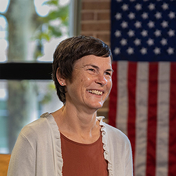 Person smiling in front of an American flag and brick wall.
