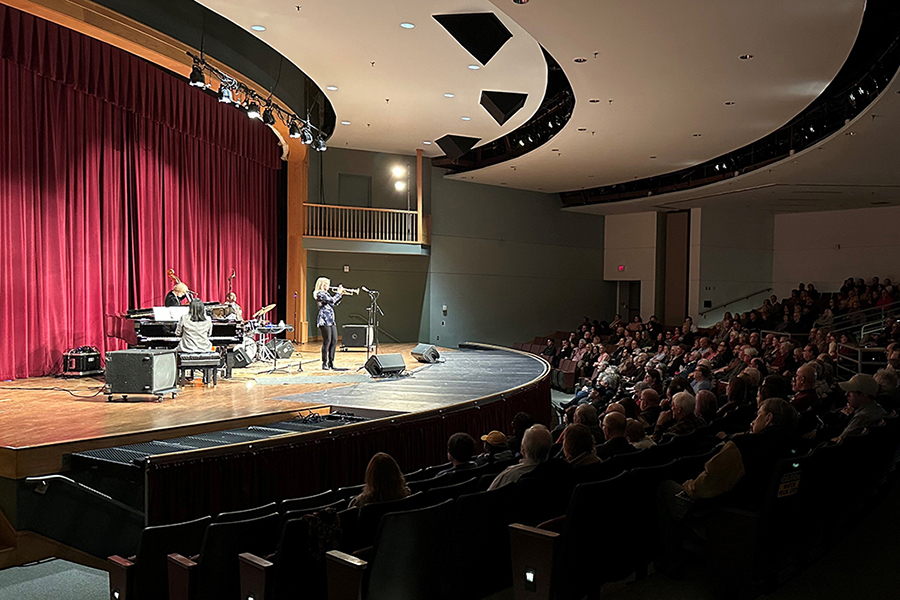 A jazz band performs on a stage with a large red curtain background before a seated audience in a darkened theater. A woman plays the trumpet in the spotlight, accompanied by a pianist, bassist, and drummer.
