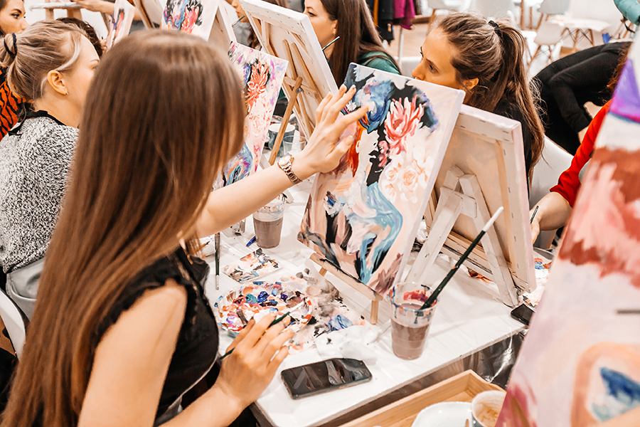 Several students are seated at a long table, each working on their own vibrant floral painting at an easel.