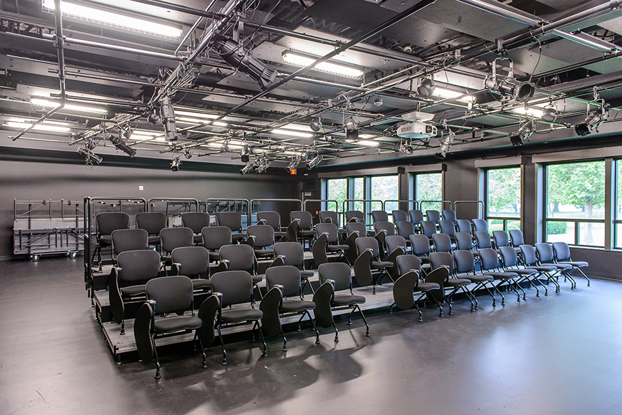 A modern studio theatre features tiered rows of black cushioned chairs facing an open floor space. The ceiling is equipped with a complex grid of professional stage lighting and a projector, while large windows along the side wall let in natural light.
