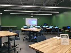 A makerspace or media lab with green walls. The room contains large wooden workbenches in the foreground and a row of computer workstations against the back wall with a projection screen.