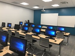 A computer lab with multiple rows of desktop monitors on long tables. The room features a dark teal accent wall with two whiteboards and a ceiling-mounted projector.