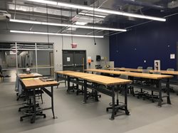 A high-ceilinged trades lab with long, heavy-duty wooden workbenches. The room has industrial lighting, a navy blue accent wall, and a large glass entryway.