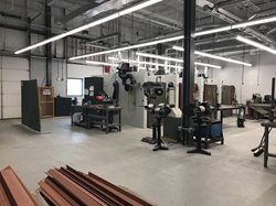 A spacious welding lab with concrete floors, featuring industrial welding booths, ventilation systems, and metal stock organized in the foreground.